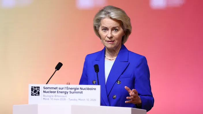 Une femme blonde aux cheveux courts parle derrière un pupitre lors d’une conférence. Elle porte une veste bleue et fait un geste de la main en s’exprimant. Il s’agit d’Ursula von der Leyen, debout devant un fond rouge, avec un micro et un panneau devant elle.