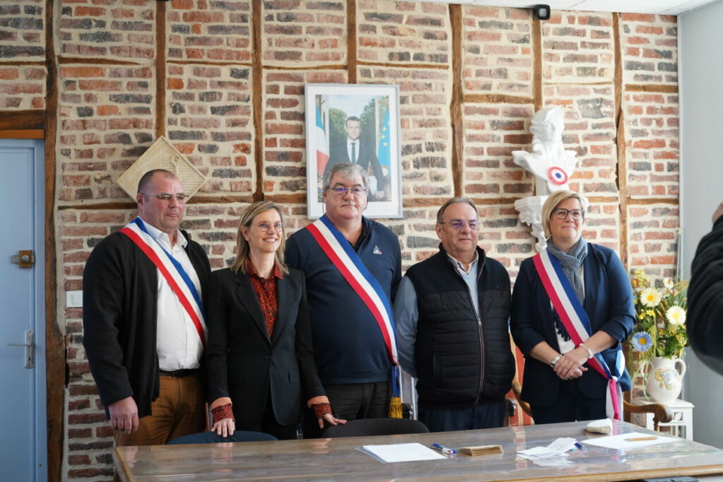 Cinq personnes posent dans une salle de mairie devant un mur en briques. Trois portent une écharpe tricolore d’élus. Au centre, une femme blonde, Agnès Pannier-Runacher en veste noire sourit. Au-dessus d’eux, un portrait officiel du président de la République est accroché au mur. Devant, une table avec des papiers et des stylos suggère une cérémonie officielle, comme une signature ou une installation d’élus.