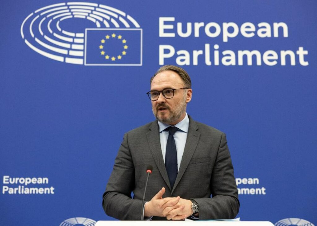 Un homme avec des lunettes et une barbe courte parle derrière un pupitre. Il porte un costume gris avec une chemise claire et une cravate sombre. Il se tient devant un fond bleu avec le logo du Parlement européen, lors d’une prise de parole officielle. Il s’agit de Dan Jørgensen.