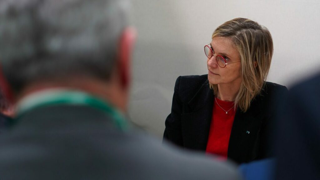Agnès Pannier-Runacher est assise lors d’une réunion, écoutant attentivement un interlocuteur hors champ. Elle porte une veste noire sur un haut rouge et des lunettes rondes rouges.